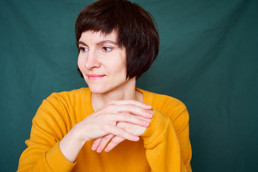 Smiling mature brunette woman looking away playful and defiant, close-up face portrait of middle-aged lady in bright yellow sweater on bright colorful dark green textile background. Generation X, 40s