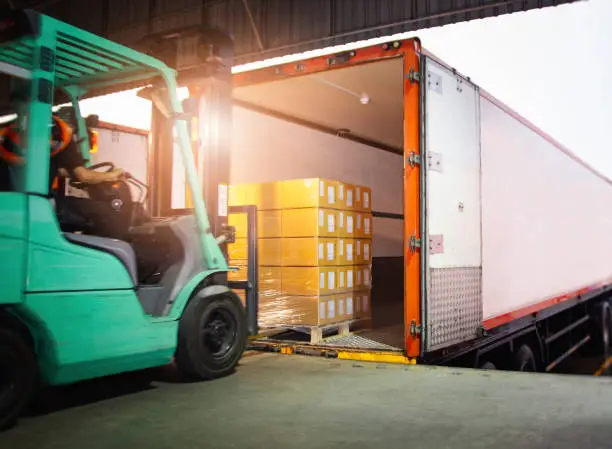 Photo of Forklift Driver Loading Package Boxes into Cargo Container. Cargo Trailer Truck Parked Loading at Dock Warehouse. Shipment Delivery Service. Shipping Warehouse Logistics. Freight Truck Transportation.