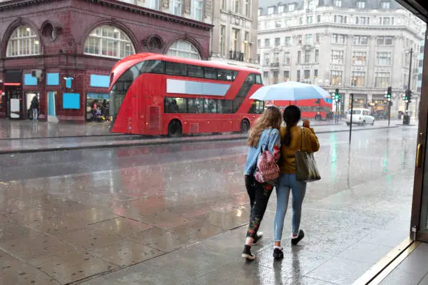 Rear view of two young women under one umbrella rushing down the street in London downtown on a rainy day, double-decker busses and cars in wet street