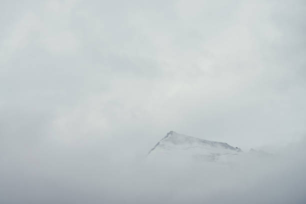 Photo of Minimal mountain landscape with high pointy rock in clouds. Minimalist mountain scenery with sharp snowy mountain peak over clouds. Snow-white pointed pinnacle above white clouds. Big top in dense fog