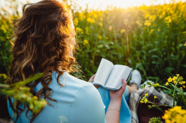 uma garota se senta em um campo em um gramado de flores amarelas, lendo um livro em um dia ensolarado de verão. uma linda garota de vestido azul lê uma bíblia. leitura aberta. lugar para texto. - lugar de devoção religiosa - fotografias e filmes do acervo