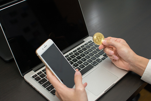 A young man holds in his right hand Bitcoin earned in virtual currency using a laptop and smartphone.