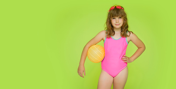 Mockup children's swimsuit. A beautiful little pre-adolescent girl in a pink swimsuit on a green background looks at the camera. Cute baby with swimming goggles and beach volleyball. Copyspace