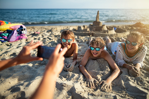Three kids having fun on the beach. They have built a sandcastle and now they are lying on sand. Mother is taking photos of the kids.\nNikon D850