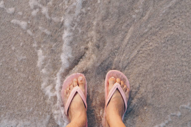 女性の足は、海水スプラッシュの背景を持つ砂の熱帯のビーチに立ってスリッパを着用しています。 - flip flop human foot sand women ストックフォトと画像