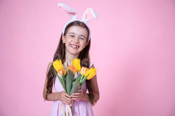 Funny little girl with easter bunny ears holding a bouquet of spring flowers copy space. Cheerful little girl with Easter bunny ears smiles and holds a bouquet of tulips in her hands on a pink studio background. holding child flower april stock pictures, royalty-free photos & images
