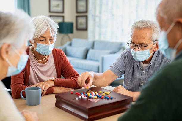 Senior friends playing chinese checkers at nursing home Group of seniors men and women wearing protective face mask playing chinese checkers at nursing home during pandemic lockdown. Old man wearing eyeglasses and surgical face mask for safety against covid-19 playing with a group of friends. Senior people enjoy a board game at care facility centre. chinese checkers stock pictures, royalty-free photos & images