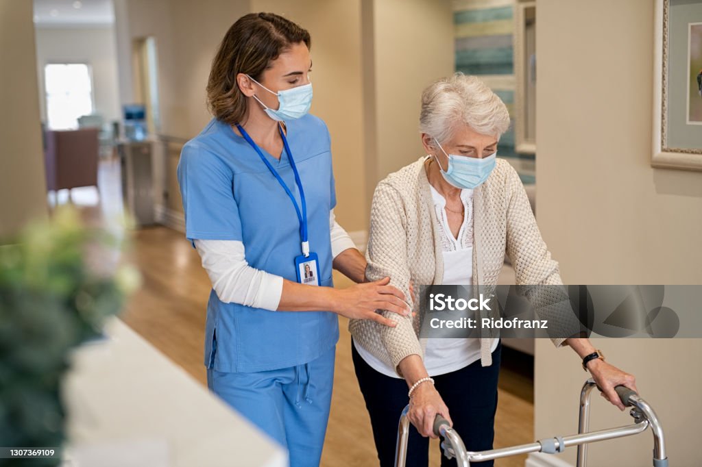 Nurse helping senior woman walk at nursing home Smiling nurse with face mask helping senior woman to walk around the nursing home with walker. Young lovely nurse helping old woman with surgical mask for safety against covid-19 using a walking frame in hospital corridor. Friendly caregiver and elderly patient walking in hallway wearing protective face mask during covid pandemic. Nurse Stock Photo