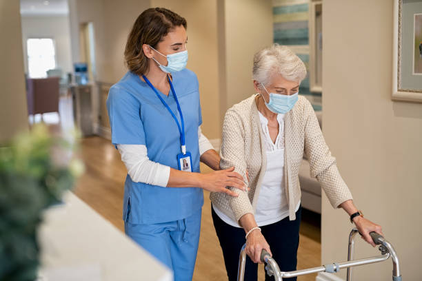 Nurse helping senior woman walk at nursing home Smiling nurse with face mask helping senior woman to walk around the nursing home with walker. Young lovely nurse helping old woman with surgical mask for safety against covid-19 using a walking frame in hospital corridor. Friendly caregiver and elderly patient walking in hallway wearing protective face mask during covid pandemic. mobility walker stock pictures, royalty-free photos & images