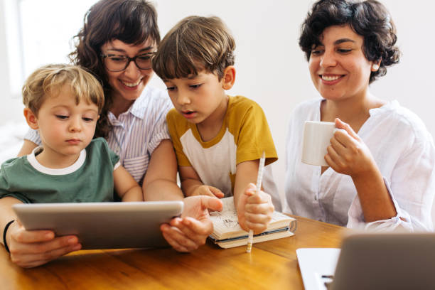 Young family at home with digital tablet Family sitting together at table using digital tablet. Woman with kids looking at laptop with her partner drinking coffee and smiling. lesbian stock pictures, royalty-free photos & images