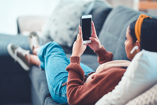Shot of a young woman using a smartphone and wireless earbuds on the sofa at home