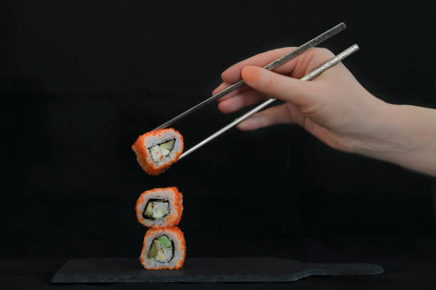 Sushi rolls lie on a tray and a hand with chopsticks reaches out to them on black background stock photo