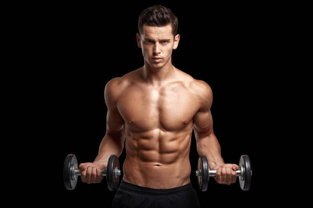 Front image of a fit, strong athletic young man with bare torso, training with dumb-bell, isolated black background. stock photo