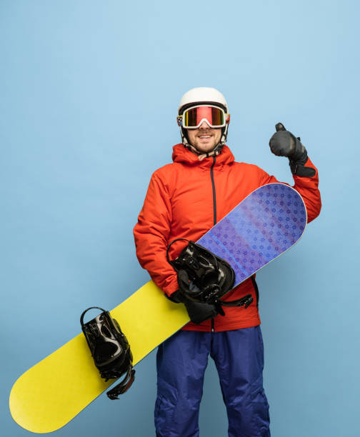 istock Young man in bright ski suit and snowboard posing isolated on blue background. 1307378548