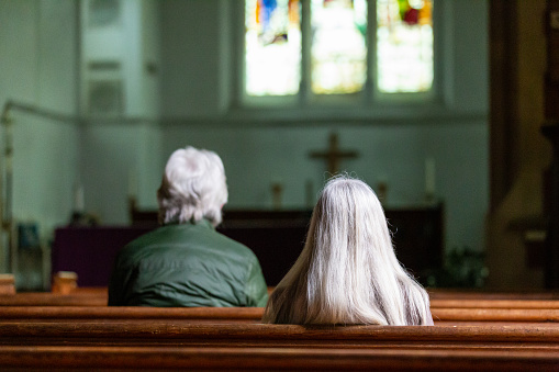 Color image depicting a rear view of two senior adults praying inside an Anglican church. Focus is on the people's heads while the altar and stained glass window is defocused beyond. Room for copy space.