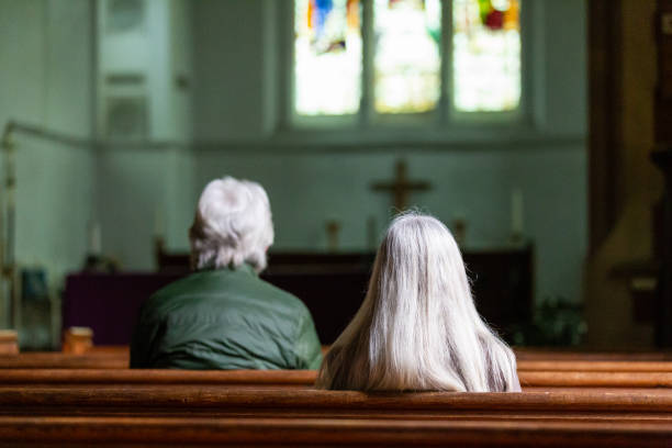 Rear view of two people praying in church Color image depicting a rear view of two senior adults praying inside an Anglican church. Focus is on the people's heads while the altar and stained glass window is defocused beyond. Room for copy space. pew stock pictures, royalty-free photos & images