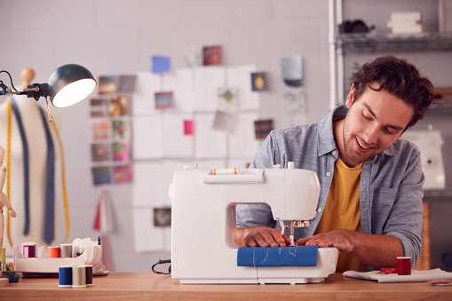 Smiling Male Student Or Business Owner Working In Fashion Using Sewing Machine In Studio