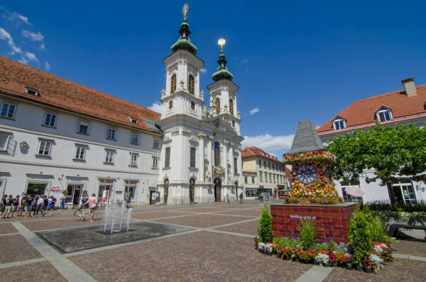 the famous clock tower, one of the city main attractions, made of flowers and mariahilfer church and square in sunny day, styria region, austria - graz austria clock tower styria imagens e fotografias de stock