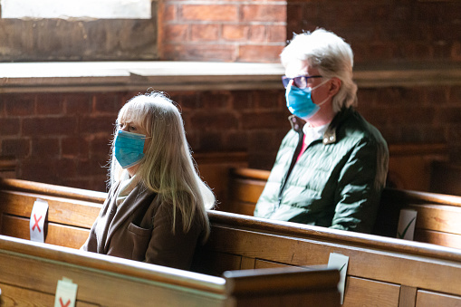 Selective focus image depicting two senior adults praying inside an Anglican church while wearing a protective face mask during the covid-19 pandemic. There are ticks and crosses on the wooden pews, indicating a social distancing system. Room for copy space.