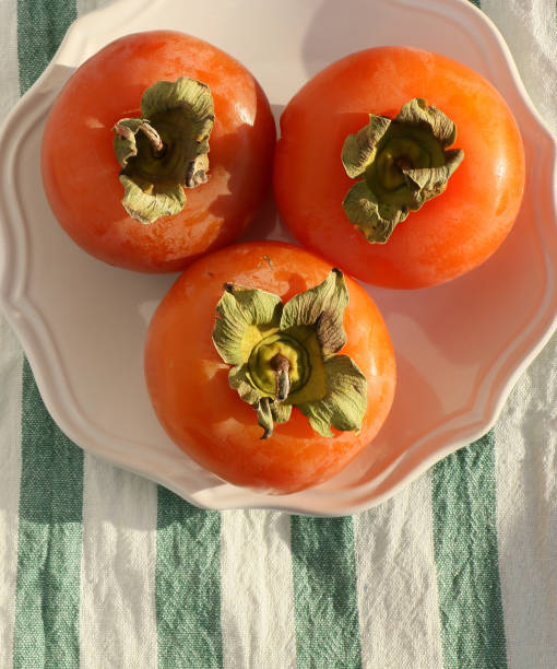 istock Fresh ripe persimmons on a white plate with a classic design on a green-white striped tablecloth, Paradise Palms 1307389744