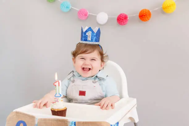 Happy smiling cute Caucasian baby boy in blue crown celebrating his first birthday at home. Child kid toddler sitting in high chair eating tasty cupcake dessert with lit candle. Happy birthday concept