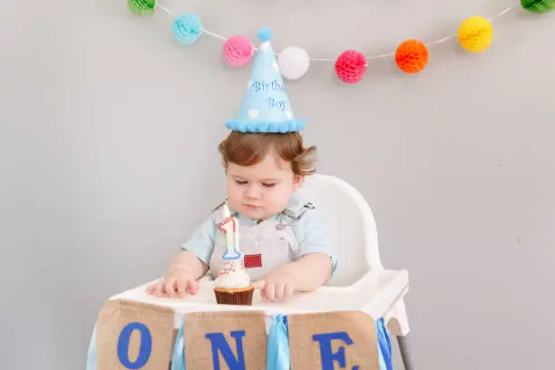 Cute adorable Caucasian baby boy in blue hat celebrating his first birthday at home. Child kid toddler sitting in high chair looking at cupcake dessert with lit candle. Happy birthday concept.