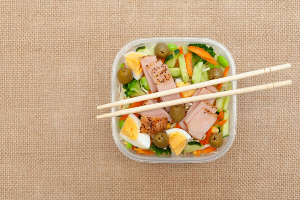 istock Healthy chicken salad with vegetables in plastic container with chopstick. Isolated on jute background. Top view. Copy space 1307391478