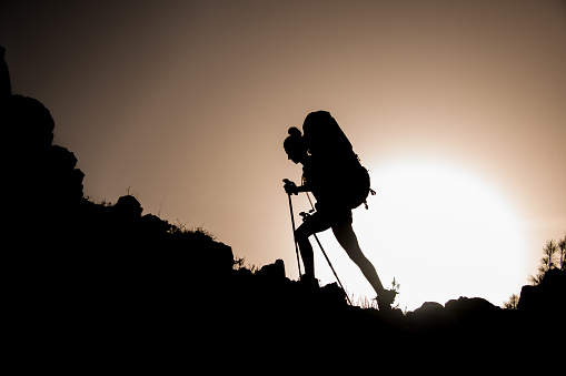 Dark silhouette of woman tourist with trekking sticks and travel backpack on her back who walks along rocky path against sky