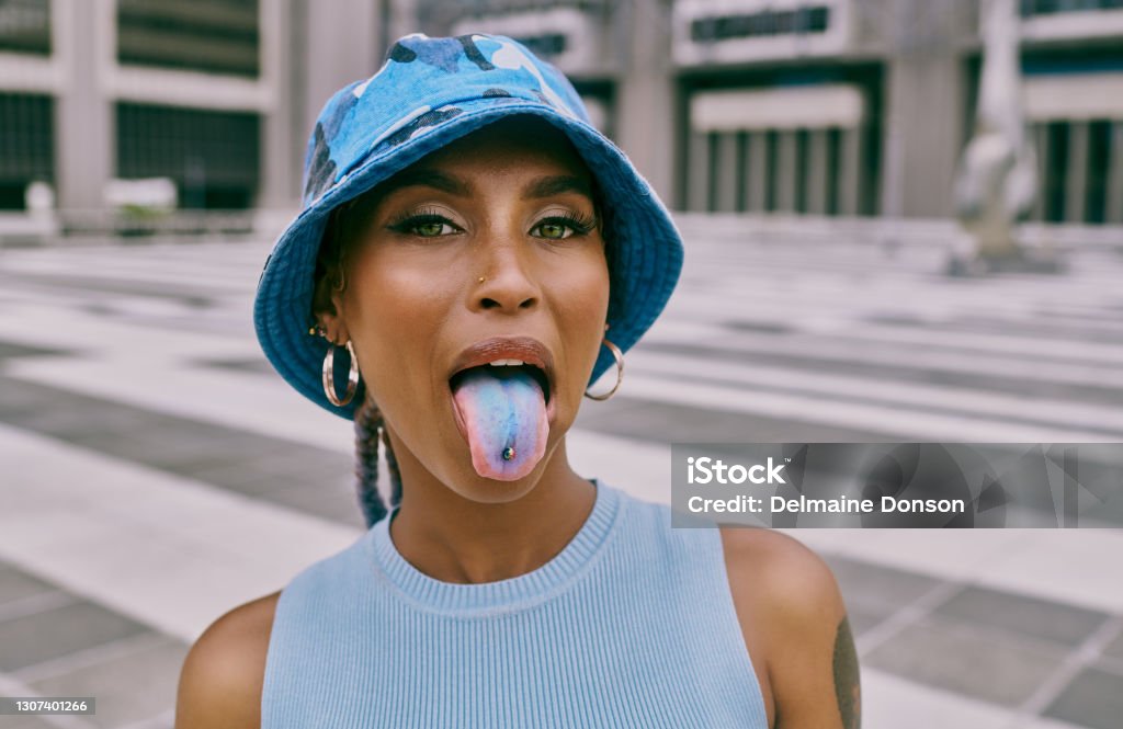 Whatever your personality, make sure it pops Shot of a trendy young woman sticking out her blue coloured tongue and showing her piercing against an urban background Sticking Out Tongue Stock Photo