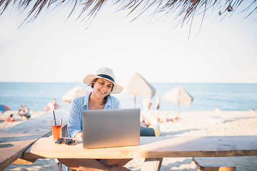 A shot of an young business woman drinking coctail and using a laptop on the beach.