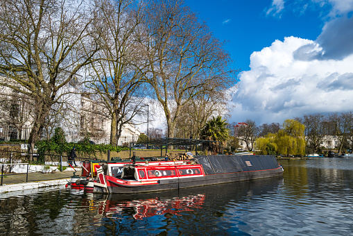 London, UK - 13 March, 2021: color image depicting houseboats and narrow boats on Regent's Canal in the Little Venice area of London, UK. Little Venice is a district in West London, England, around the junction of the Paddington Arm of the Grand Union Canal, the Regent's Canal, and the entrance to Paddington Basin. People are walking and joggers jogging on the side of the canal.