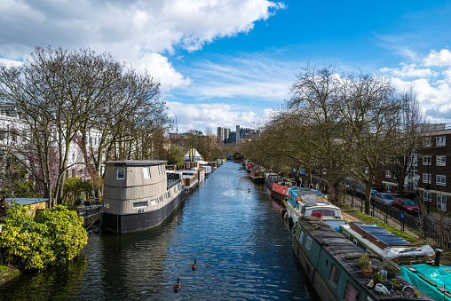 Color image depicting houseboats and narrow boats on Regent's Canal in the Little Venice area of London, UK. Little Venice is a district in West London, England, around the junction of the Paddington Arm of the Grand Union Canal, the Regent's Canal, and the entrance to Paddington Basin. People are walking and joggers jogging on the side of the canal.