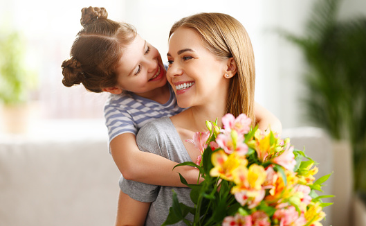 Cheerful kid embracing woman sitting on sofa with bouquet of Alstroemeria flowers on Mothers Day