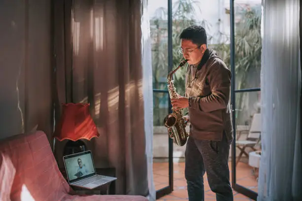 Lifehack asian chinese mid adult man practicing saxophone at living room online virtual class with his tutor using laptop