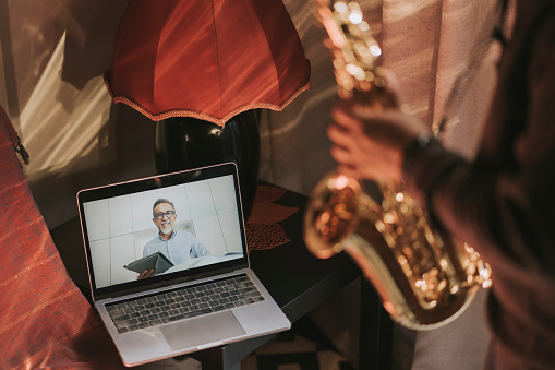 Lifehack asian chinese mid adult man practicing saxophone at living room online virtual class with his tutor using laptop