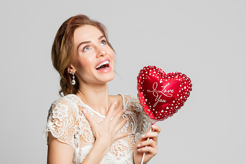 Happy blond woman wearing weeding dress, holding red heart shape balloon. Studio shot against grey background.