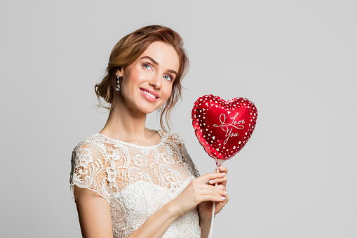 Happy blond woman wearing weeding dress, holding red heart shape balloon. Studio shot against grey background.