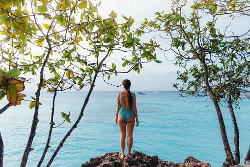 Woman with long hair in blue swimsuit enjoying the scenic summer sunset looking at the pure crystal clean Indian Ocean staying on the shore between trees on Zanzibar, Tanzania
