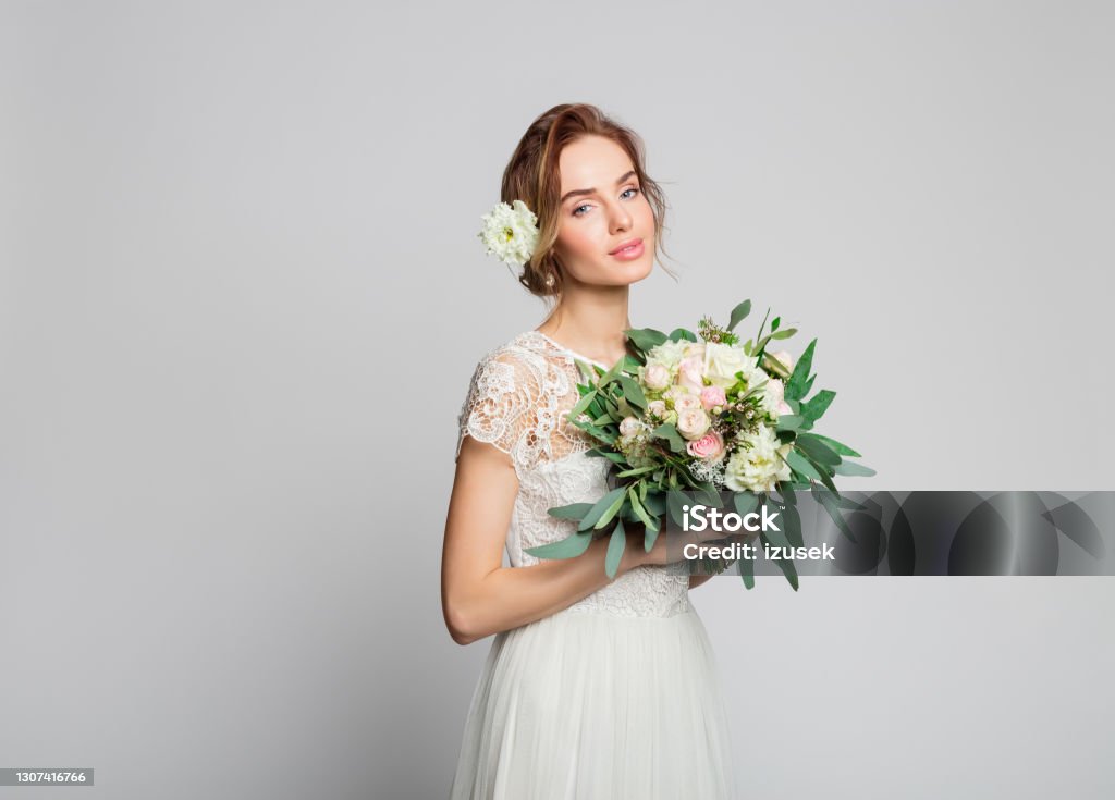 Portrait of beautiful bride Dreamy blond woman wearing weeding dress and holding bouquet. Studio shot against grey background. Luxury Stock Photo