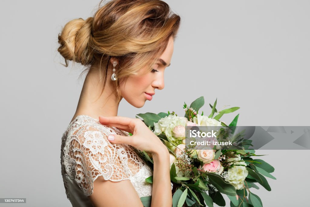 Portrait of beautiful bride Side view of blond woman wearing weeding dress and holding bouquet. Studio shot against grey background. Blond Hair Stock Photo