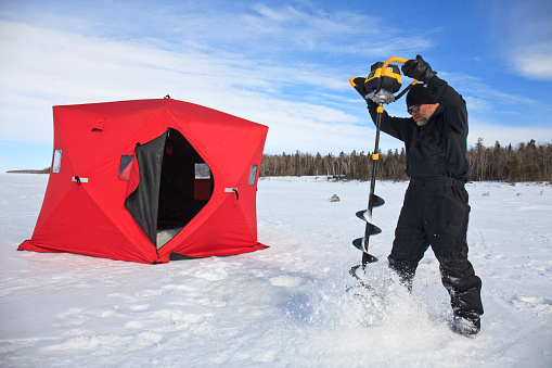 A caucasian male drilling a hole with an ice auger for ice fishing expedition in Manitoba, Canada. A red collapsible ice fishing shelter is also in the image.