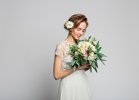 Dreamy blond woman wearing weeding dress and holding bouquet. Studio shot against grey background.