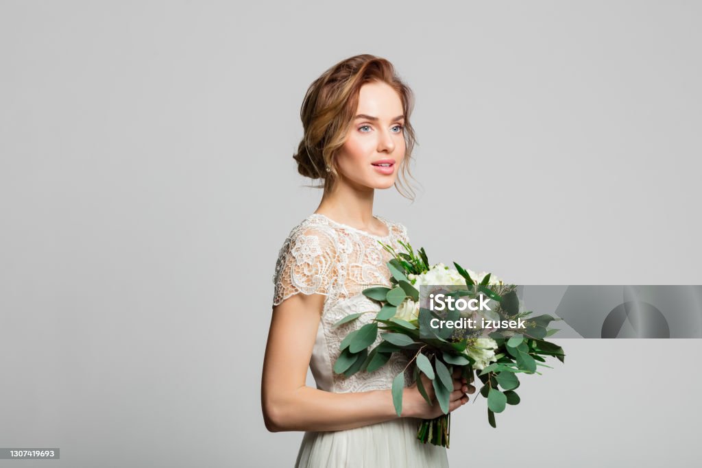 Portrait of beautiful bride Dreamy blond woman wearing weeding dress and holding bouquet, looking away. Studio shot against grey background. Adult Stock Photo