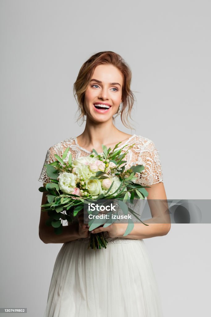 Portrait of beautiful bride holding flowers Happy blond woman wearing weeding dress and holding bouquet. Studio shot against grey background. Adult Stock Photo