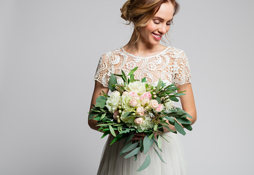 Happy blond woman wearing weeding dress and holding bouquet. Studio shot against grey background. Close up of flowers.