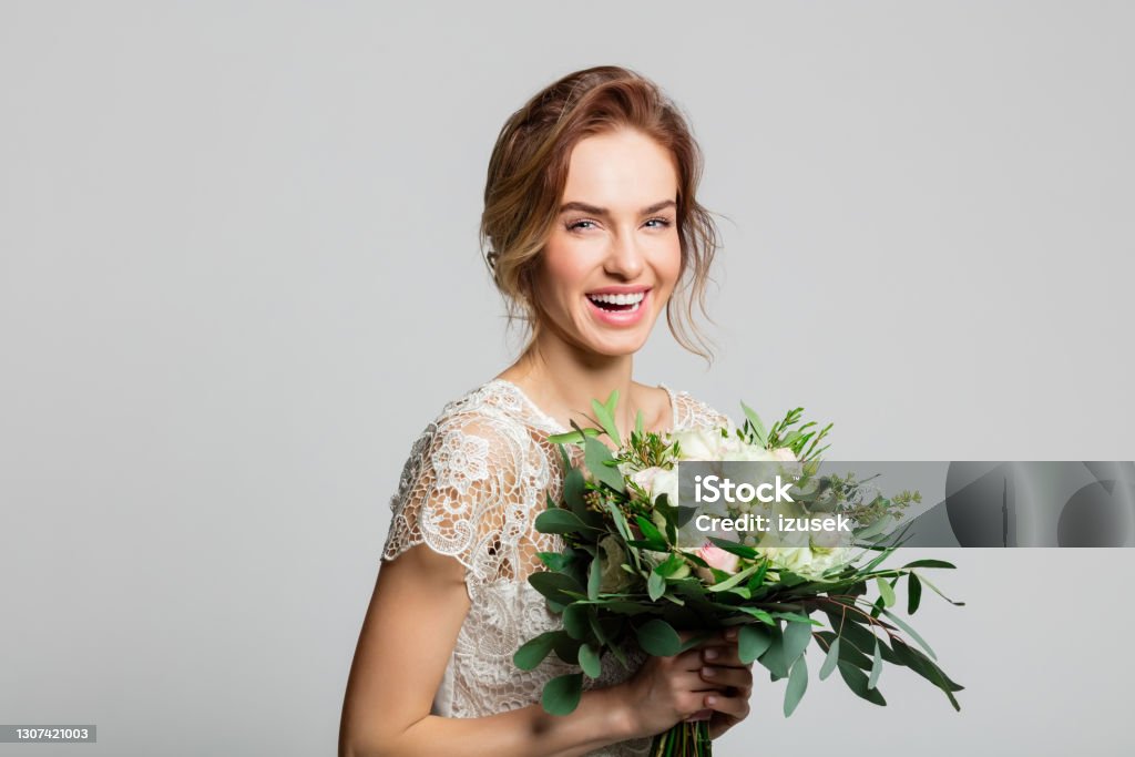 Portrait of beautiful bride holding flowers Cheerful blond woman wearing weeding dress and holding bouquet, laughing at camera. Studio shot against grey background. Adult Stock Photo