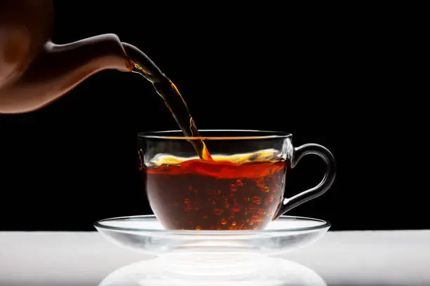 Pouring black tea into transparent cup isolated on black background