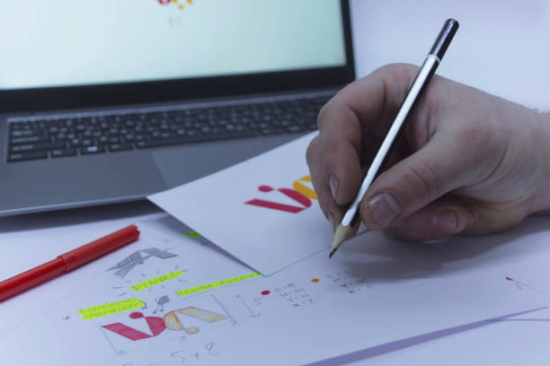 istock Creative workplace of a Graphic Designer. A man in the office is developing a logo on the table against the background of printed sketches and a laptop. 1307423047