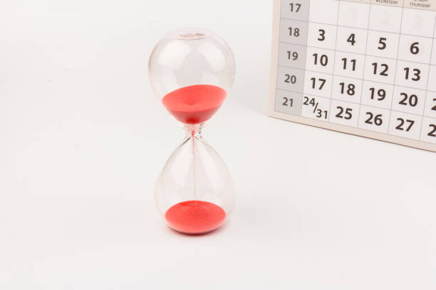 istock Time passing concept. Crystal hourglass with red sand and calendar on light background 1307423435