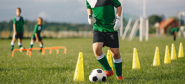 Kid soccer player dribbling through cones. Boy in soccer uniform practice with ball. Child kicking ball on grass. Young athlete improving football dribbling skills